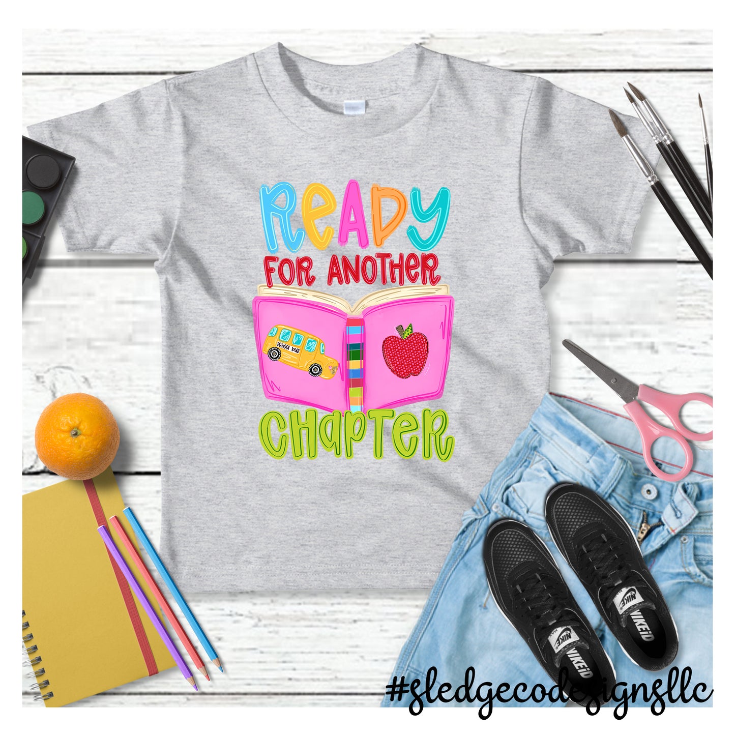 Ready for Another Chapter Teacher Shirt featuring an open book, school bus, and apple on a custom unisex T-shirt by Sledge Co Designs.